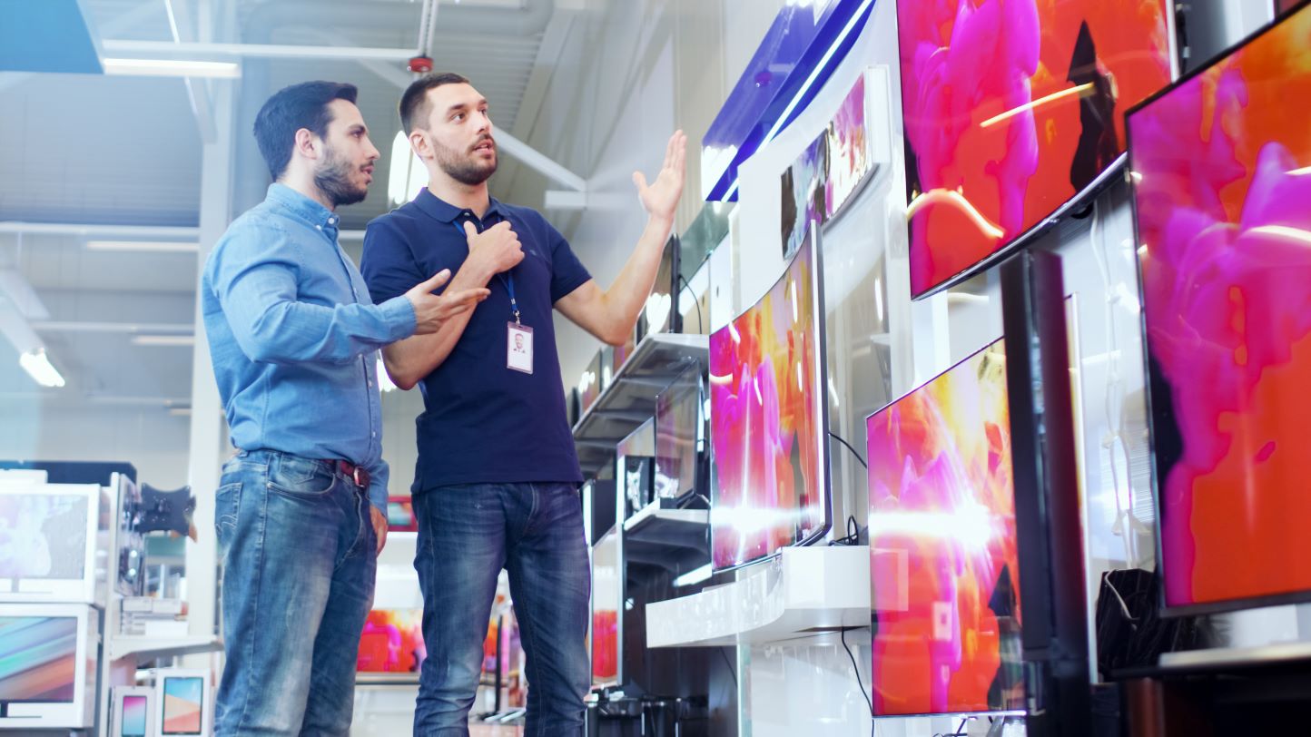 In the Electronics Store Professional Consultant Shows Latest 4K UHD TV's to a Young Man, They Talk about Specifications and What Model is Best for Young Man's Home. Store is Bright, Modern and Has all the Latest Models.
