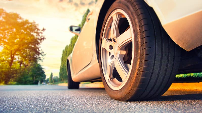Close-up of a car tire on a silver vehicle, parked on a paved road with a scenic background of trees and a warm sunset