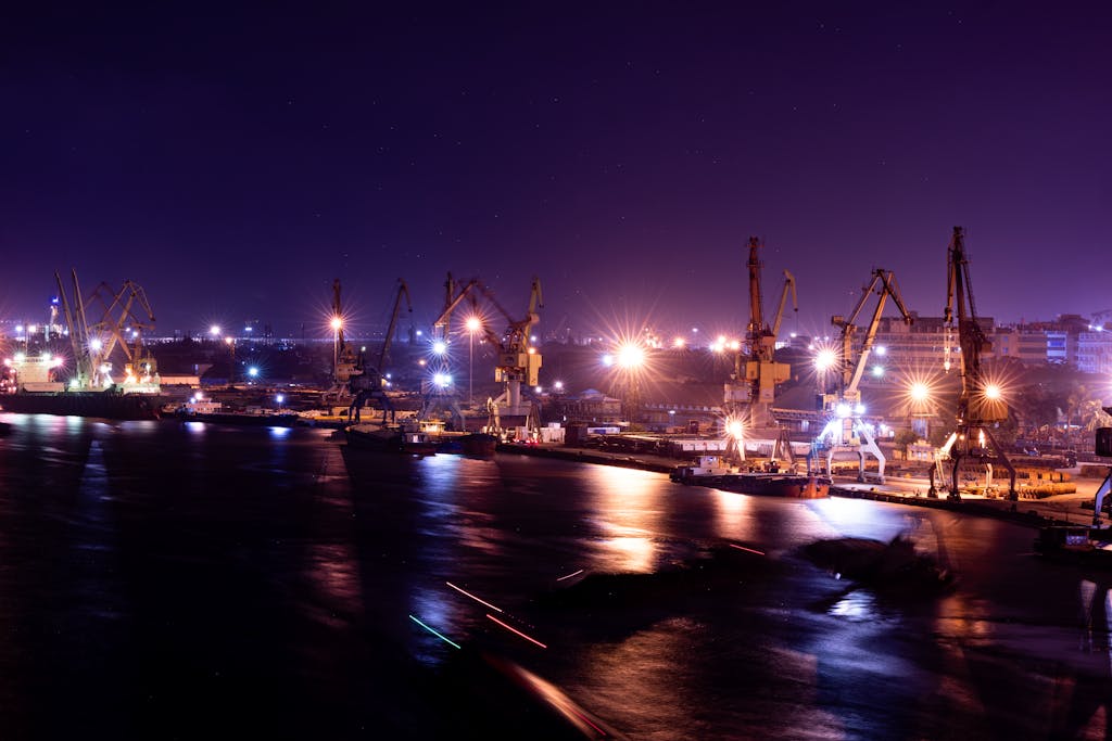 A serene night view of illuminated shipping cranes at a bustling harbor, reflecting lights over calm water.