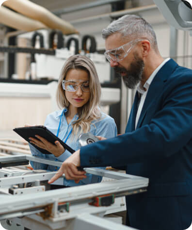bearded man in safety glasses pointing into a machine while a women in safety glasses takes notes on an ipad