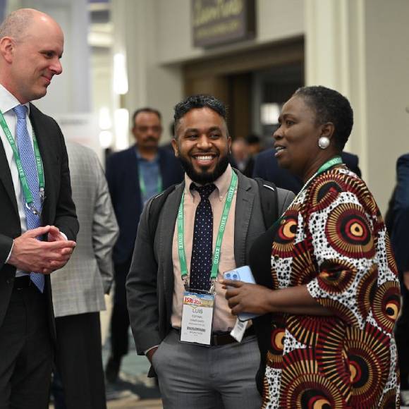 A group of three diverse professionals engaged in conversation at a conference. They are wearing business attire with green lanyards and name badges, smiling and interacting in a lively networking environment with other attendees in the background.