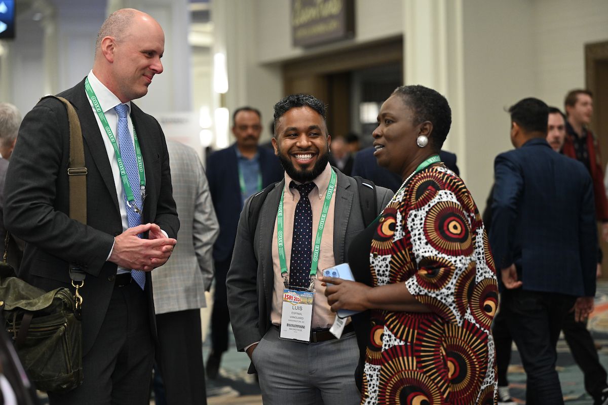A group of three diverse professionals engaged in conversation at a conference. They are wearing business attire with green lanyards and name badges, smiling and interacting in a lively networking environment with other attendees in the background.