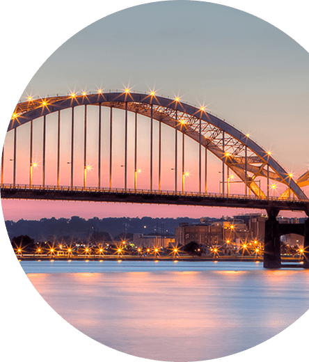 A beautifully lit arch bridge stretching over a calm river at sunset, with city lights reflecting on the water and a pastel-colored sky in the background.